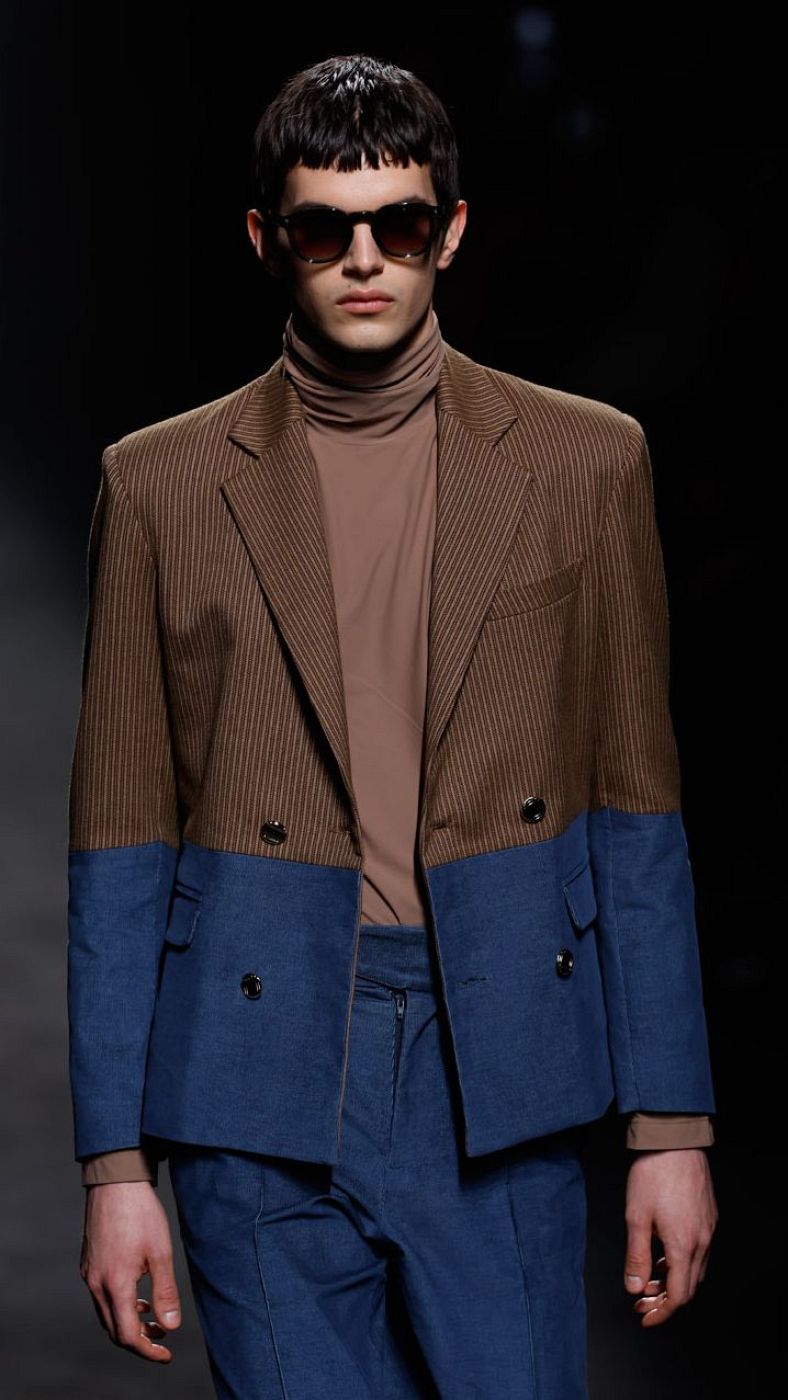 Modelo masculino con gafas de sol, presentando un diseño con chaqueta bicolor (marrón y azul marino) y jersey de cuello alto.  Silueta moderna y original.