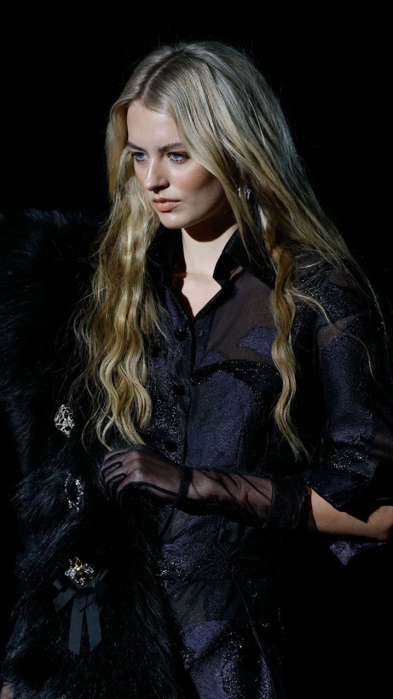 Modelo con cabello rubio y ondulado, camisa azul brillante semitransparente, guantes negros y estola negra con broches. Fondo negro.