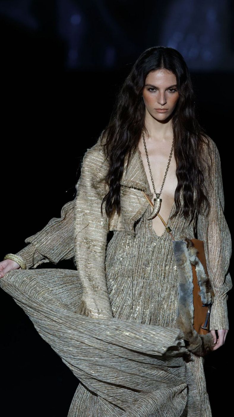 Modelo con vestido largo dorado plisado, mangas abullonadas y escote en V. Accesorios dorados y bolso marrón. Desfile de moda.