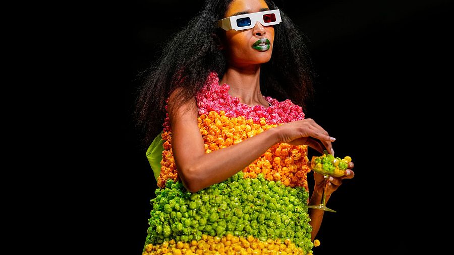 Modelo de piel oscura con vestido de palomitas de maíz, gafas 3D blancas y maquillaje vibrante desfila sobre fondo negro.
