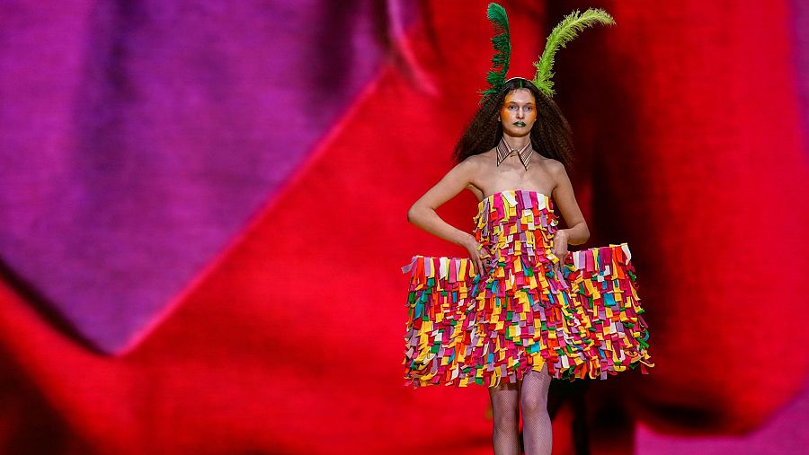 Modelo con vestido corto multicolor y tocado de plumas verdes en desfile de moda.  Maquillaje anaranjado, escenario con telón rojo y morado.