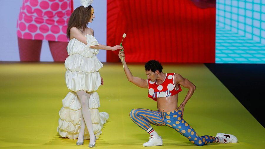 Modelo entrega flor a bailarín arrodillado en pasarela amarilla; ella viste vestido blanco con máscara dorada, él, top rojo con lunares y mallas multicolor.