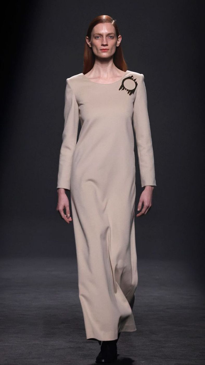 Modelo con vestido beige, manga larga y hombreras, desfilando. Broche oscuro en el pecho. Pelo rojizo recogido, expresión seria.