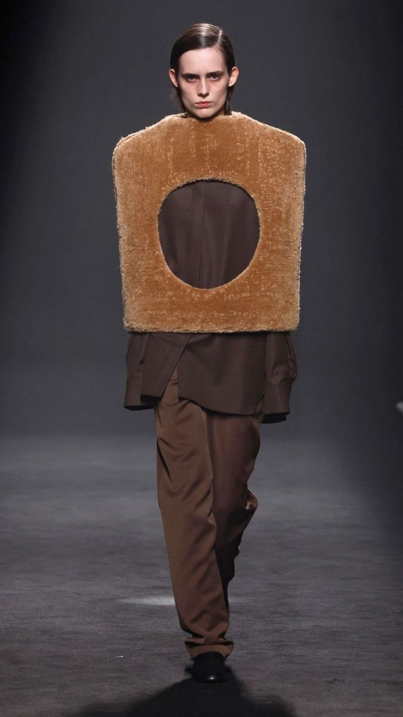 Modelo con atuendo marrón oscuro y pieza rectangular de textura afelpada marrón claro, en desfile de moda. Pelo corto y expresión seria.