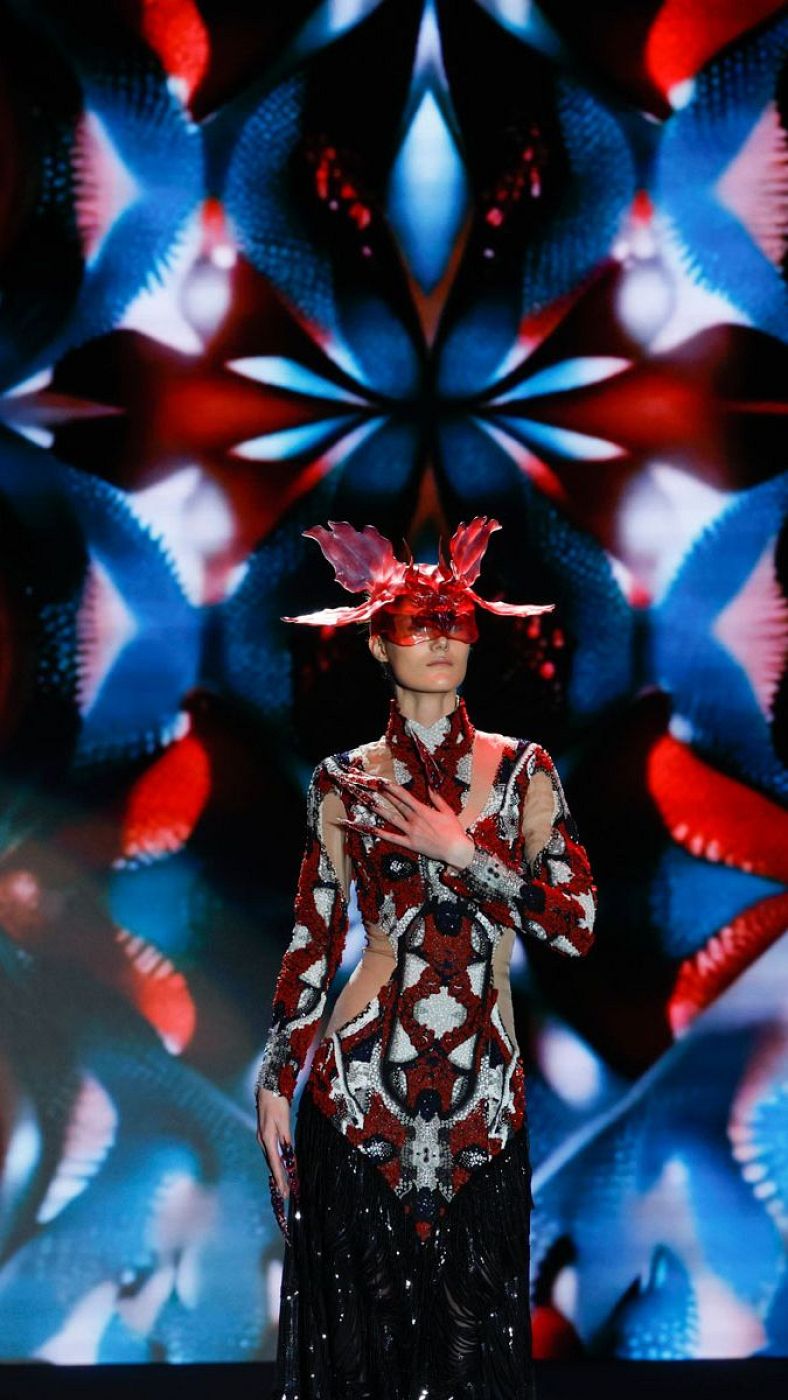 Modelo presenta diseño JNORIG: traje rojo, blanco y negro con recortes, flecos y tocado. Pasarela con pantalla de fondo rojo y azul.