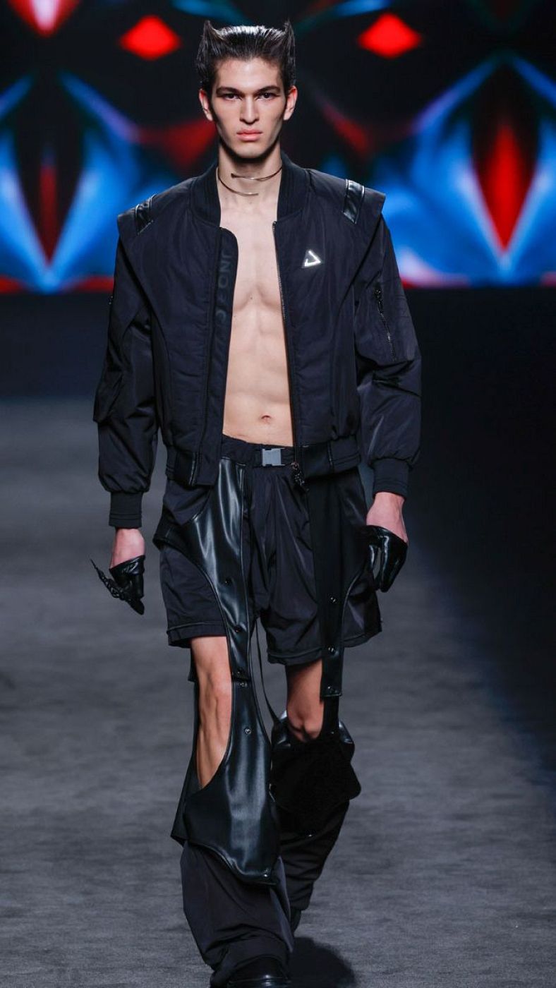 Modelo masculino desfila con un conjunto negro: bomber corta, pantalones cortos y pieza de tela con aberturas.  Guantes con detalles de garras y collar metálico completan el look.