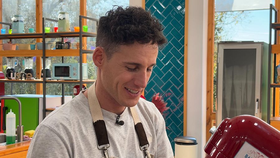 Pol Espargaró, con delantal y micrófono, en la cocina de un programa de repostería.  Ambiente colorido con utensilios pastel y azulejos verdes.