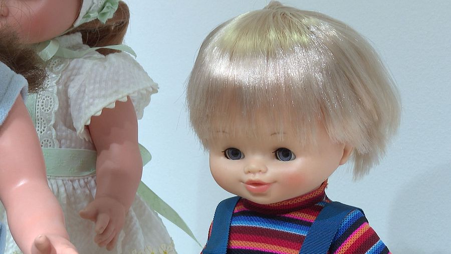 Dos muñecas, una con vestido blanco y lazo verde, la otra con jersey de rayas y peto azul, parecen caminar juntas.