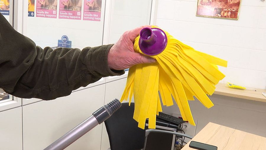 Mano sujetando fregona amarilla y mango gris; fondo con mesa, silla y teclado; posible demostración o entrevista.