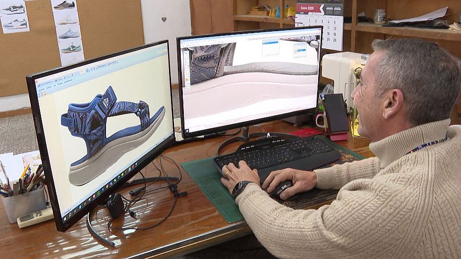 Diseñador trabajando en un modelo 3D de una sandalia azul, usando dos monitores, ratón y teclado.  Se observan bocetos y una máquina de coser.