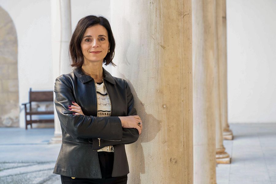 Retrato de Carmen Nestares con chaqueta negra, jersey claro y pantalones oscuros; pelo castaño corto y liso; expresión seria; brazos cruzados; fondo con columnas y suelo de baldosas.