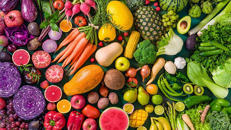 Abundancia de frutas y verduras de distintos colores y texturas, algunas enteras y otras cortadas, dispuestas en un degradado sobre fondo verde oscuro.