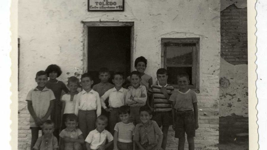 Grupo de niños posando ante una escuela rural de una planta en Toledo.  Placa cerámica con el número 23 visible sobre la puerta.  Imagen en blanco y negro.