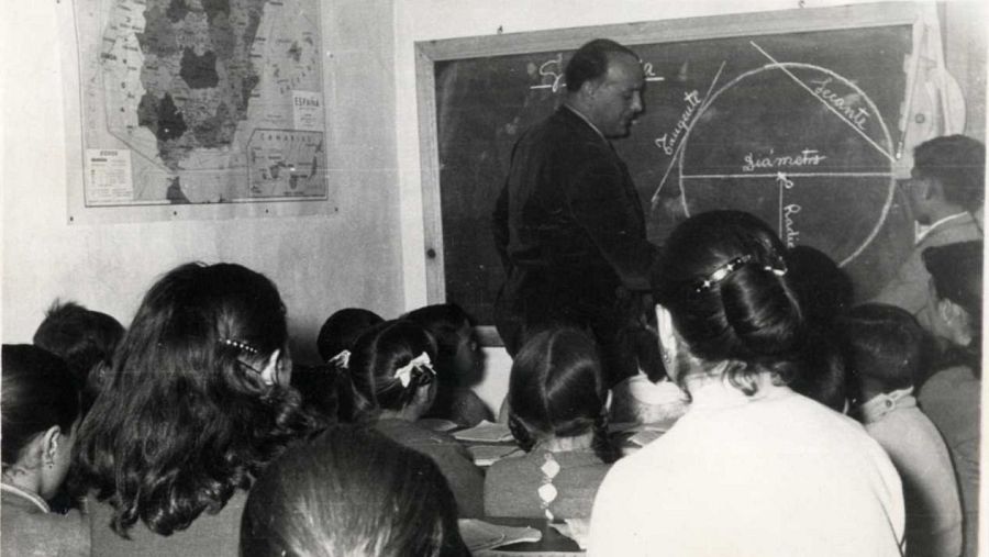 Aula de escuela rural: profesor en traje explica geometría en pizarra con figuras geométricas; alumnos atentos.