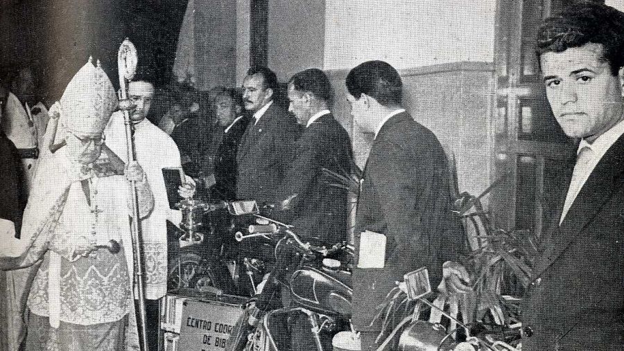 Arzobispo bendice motocicletas ante hombres en traje.  Caja con inscripción 