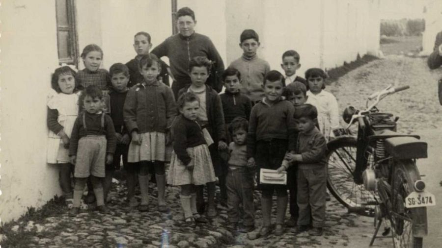 Grupo de niños, vestidos con ropa de los años 50-60, posan junto a una motocicleta clásica (matrícula SG 3474) en una calle empedrada.