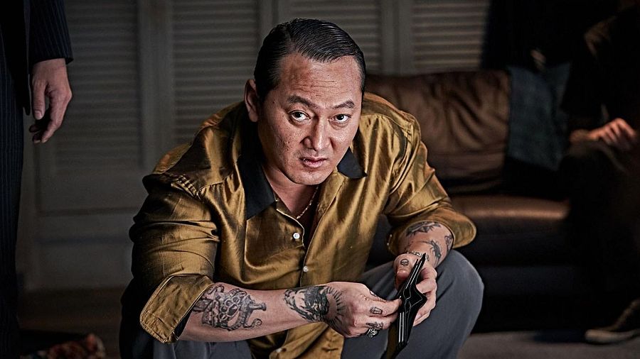 Jeong Man-shik, en primer plano, viste camisa dorada y revisa una cartera negra.  Escena oscura con atmósfera tensa; tatuajes visibles en sus brazos.