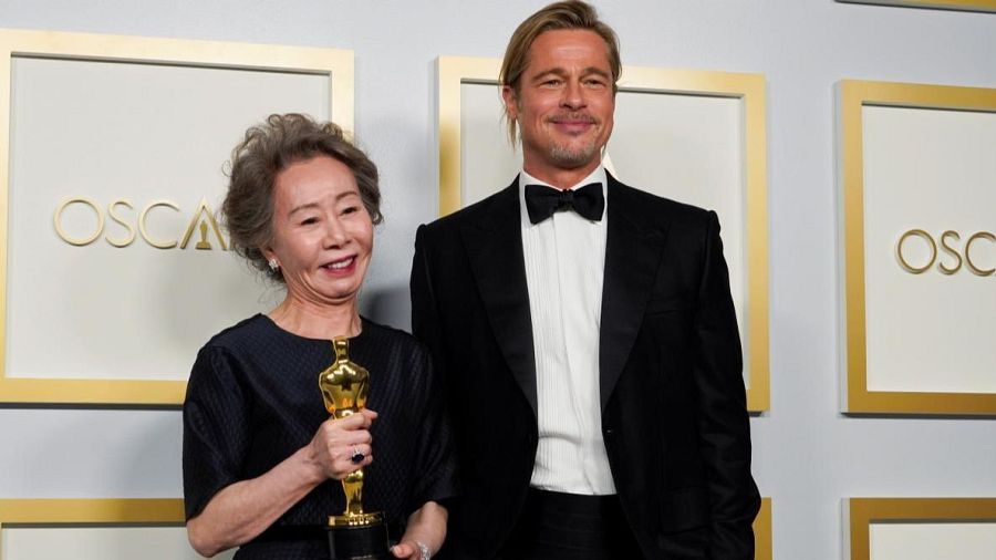 Youn Yuh-jung, con vestido oscuro, sostiene un Oscar; Brad Pitt, en esmoquin, la observa. Fondo con detalles dorados del logo de los Oscar.