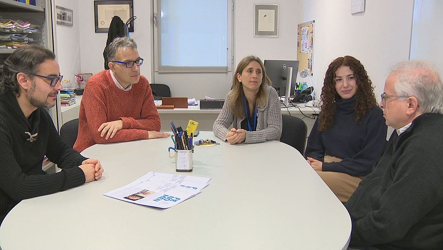 Reunión formal de cinco personas alrededor de una mesa; papeles y bolígrafos sobre la superficie.  Ambiente de oficina.