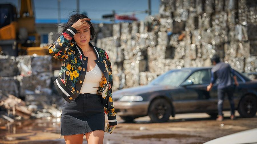 Mujer en primer plano con chaqueta bomber y minifalda, en un desguace. Al fondo, un hombre junto a un coche. Atmósfera tensa.