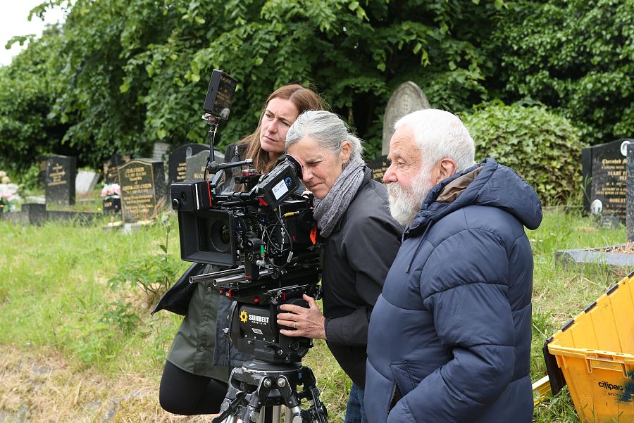 Mike Leigh observa el rodaje en un cementerio; dos mujeres operan una cámara profesional durante la grabación de una escena.