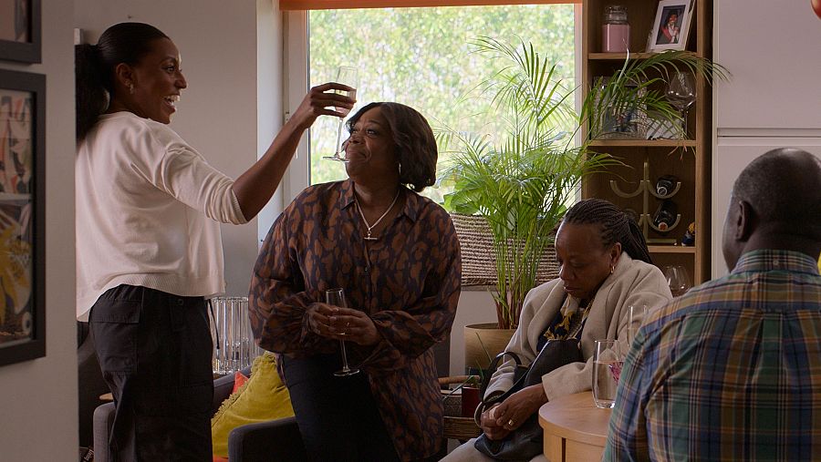 Cuatro personas afrodescendientes comparten un momento en un salón. Dos mujeres brindan con copas, mientras otras dos se sientan a una mesa en una escena cálida e iluminada.