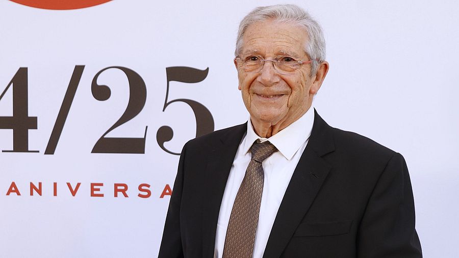 Actor en traje oscuro sonríe en evento del 25 aniversario. Fondo blanco con letras rojas y negras '4/25 ANIVERSA'.