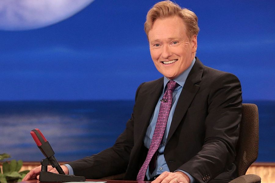 Presentador de televisión en su programa, sentado a una mesa con micrófono, traje oscuro, camisa azul y corbata morada, sonriendo a cámara. Fondo azul con imagen de mar y luna.