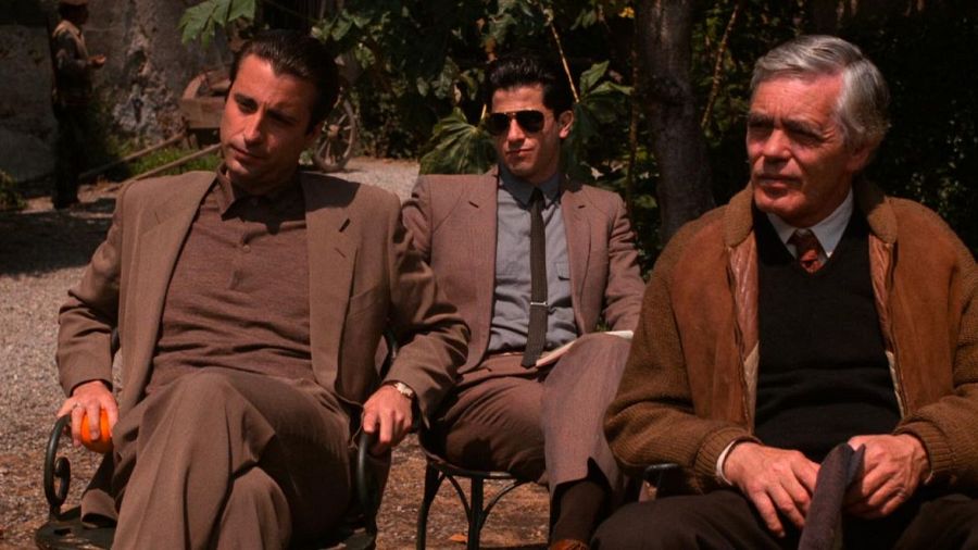 Tres hombres en trajes sentados en sillas de rejilla metálica en un jardín, uno con una fruta naranja.  Ambiente sereno y formal.