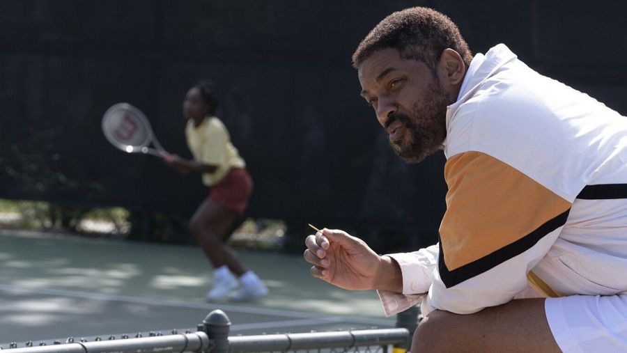 Will Smith, con chaqueta deportiva blanca y naranja, observa un partido de tenis desde el banquillo, sosteniendo un objeto en su mano.