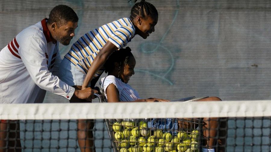 En una pista de tenis, Will Smith empuja un carrito con dos niñas sonrientes.  Fondo gris con grafitis.