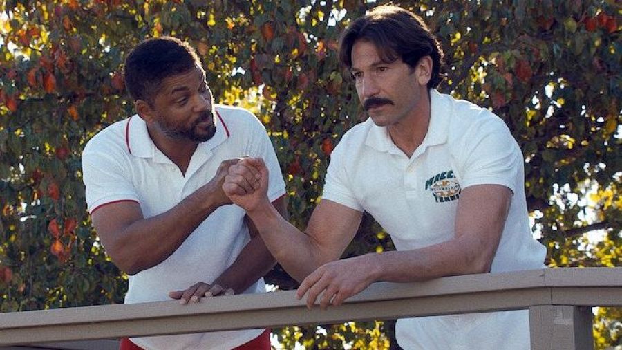 Escena de película: dos hombres con polos blancos se dan la mano en una barandilla, fondo con árboles.  Uno viste un polo con ribetes rojos; el otro, un polo con el logo 