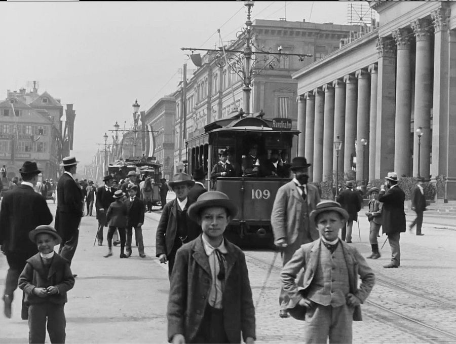 Tranvía 109 en una calle transitada de principios del siglo XX.  Gente con ropa de época y dos niños en primer plano. Imagen en blanco y negro.