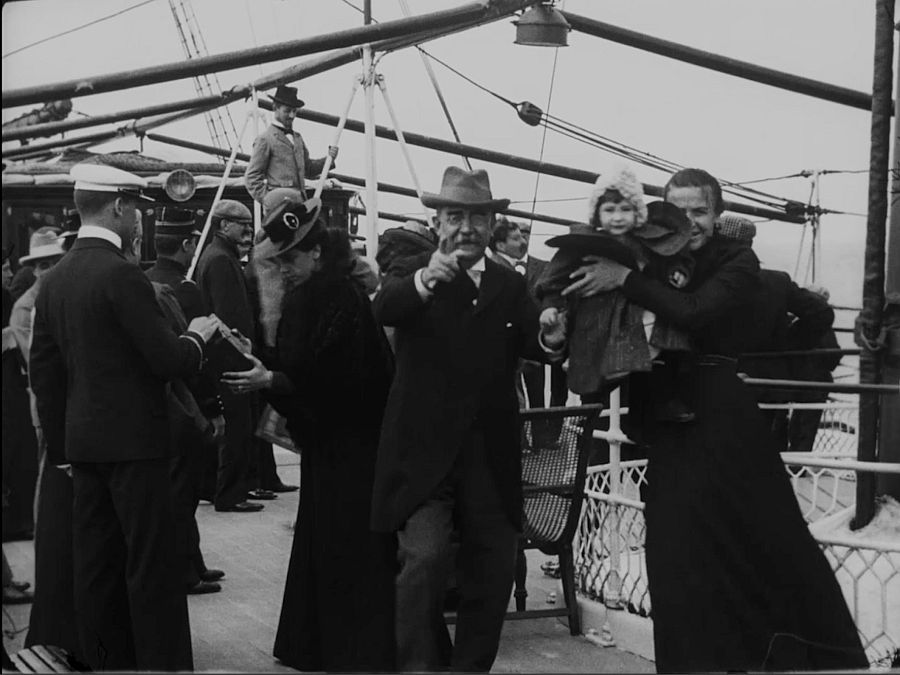 Escena en cubierta de barco: un oficial recibe una maleta; mujeres, una con niña, y otros pasajeros presentes.  Fotograma en blanco y negro con atmósfera portuaria.