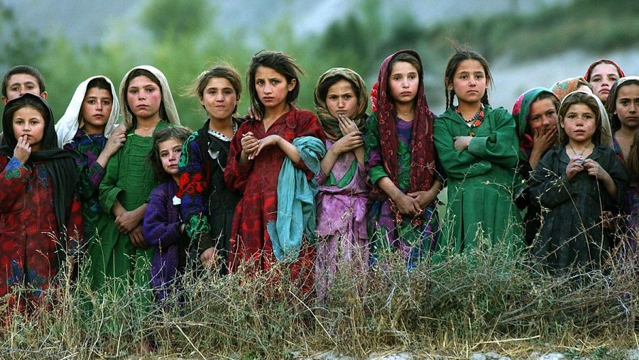 Niñas con ropas tradicionales, expresiones serias, frente a matorrales secos. Fotografía de Emilio Morenatti, tomada al atardecer, con posible trasfondo social.