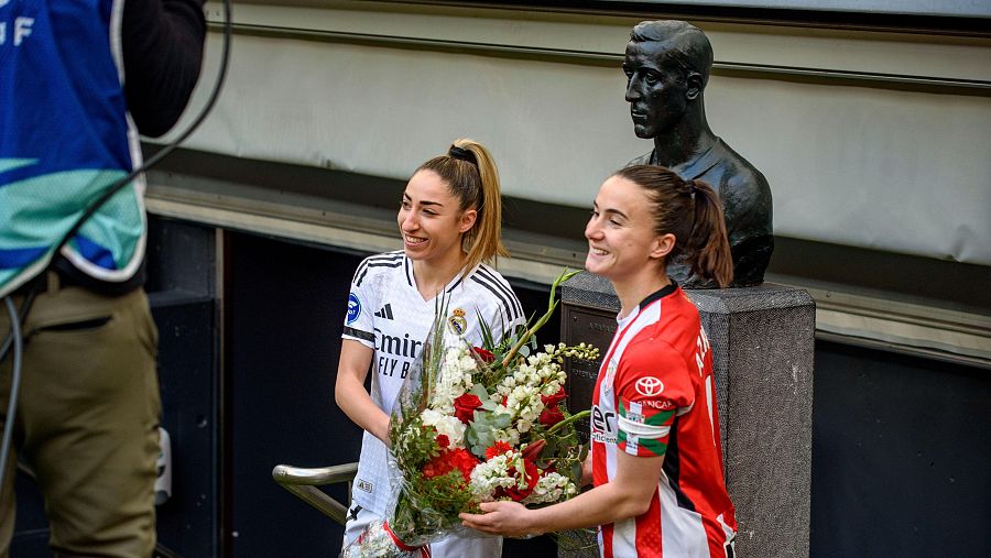 Intercambio de flores entre jugadoras de fútbol tras un partido.  Una camiseta blanca con el logo de Emirates, la otra rojiblanca con el logo de Kutxabank.  Ambas sonríen.