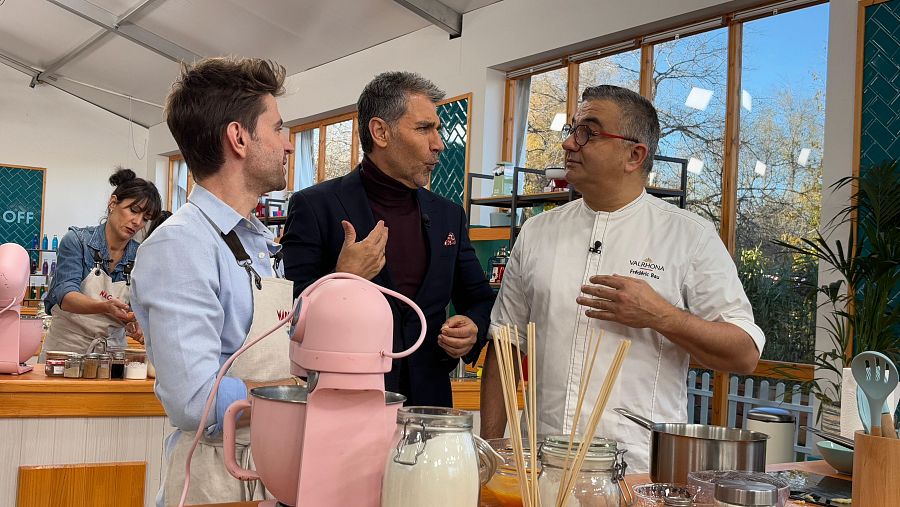 Tres chefs, en un plató de cocina, conversan. Uno lleva una chaquetilla con logo de Valrhona; hay una batidora rosa en primer plano.