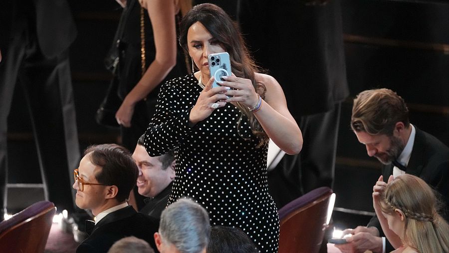 Actriz con vestido negro de lunares fotografiando con su móvil en una ceremonia formal.  Se observan otros asistentes elegantemente vestidos.