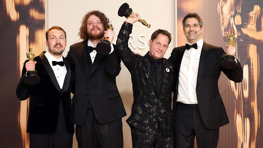 Cuatro hombres en trajes y pajaritas, sosteniendo estatuillas de los Oscar, posan en un photocall.  Uno luce chaqueta de terciopelo.