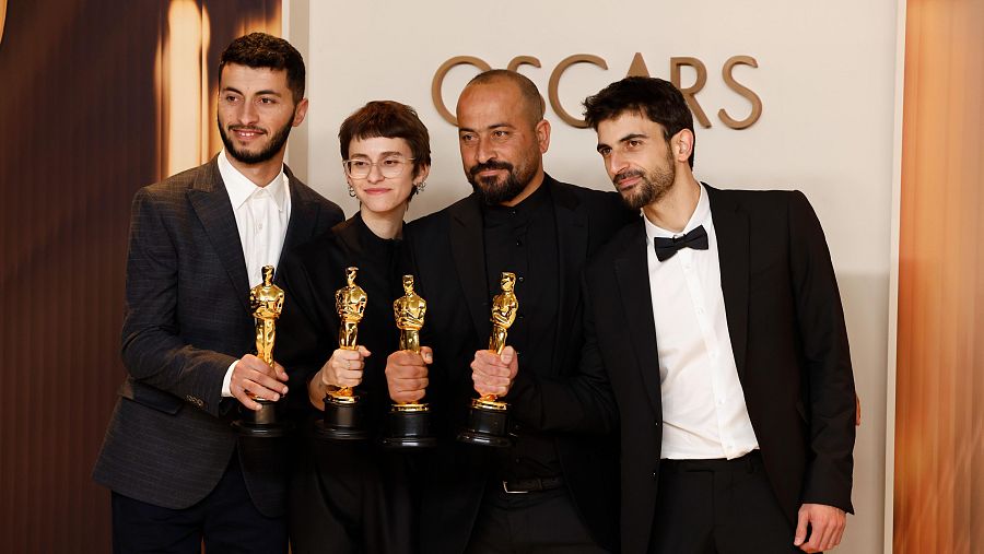 Grupo de cuatro personas en atuendos formales sosteniendo estatuillas de los Oscar, con el logo de la premiación al fondo.