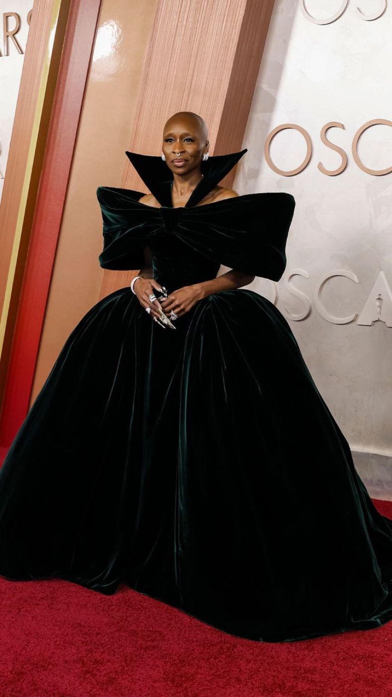 Cynthia Erivo luce un vestido verde oscuro con lazo y cuello alto en la alfombra roja.  Cabeza rapada y uñas con pedrería.