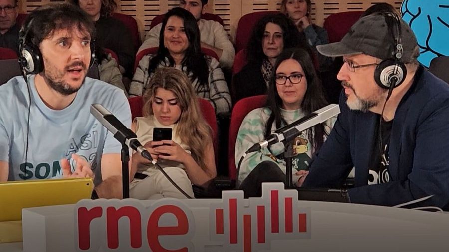 Presentadores de radio en estudio durante grabación. Uno viste camiseta con logo del programa; el otro, gorra y chaqueta. Público presente.
