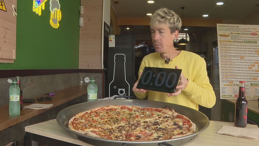 Hombre con jersey amarillo inicia un reto de comida en pizzería; cronómetro a cero, pizza grande en bandeja, bebidas en barra.