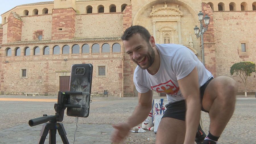 Reto en directo: hombre con camiseta blanca realiza un desafío, transmitiendo desde un teléfono en trípode en una plaza adoquinada con un muñeco de nieve y pizarra cerca.