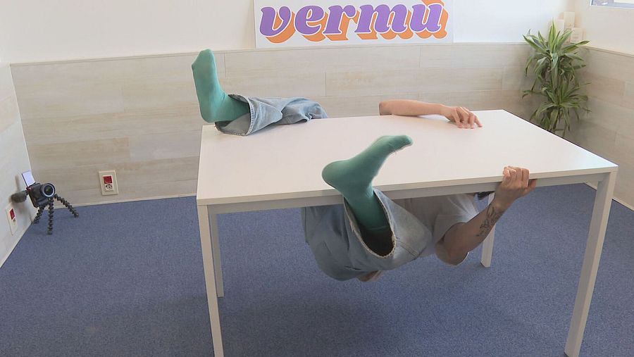 Persona intentando voltear una mesa blanca rectangular en un reto. Lleva calcetines verdes y vaqueros; se observa un tatuaje. Cámara grabando.