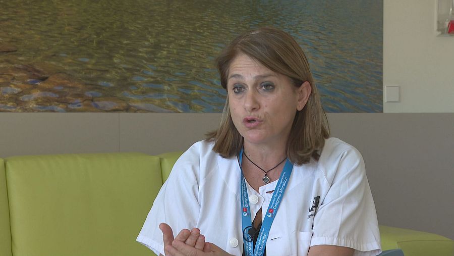 Mujer con bata blanca y acreditación hospitalaria, gesticulando. Fondo con sillón verde e imagen de paisaje.