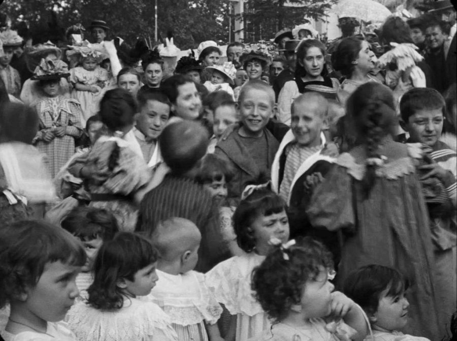 Escena bulliciosa y alegre de niños, algunos disfrazados, en un evento al aire libre.  Fotograma en blanco y negro de una película antigua.