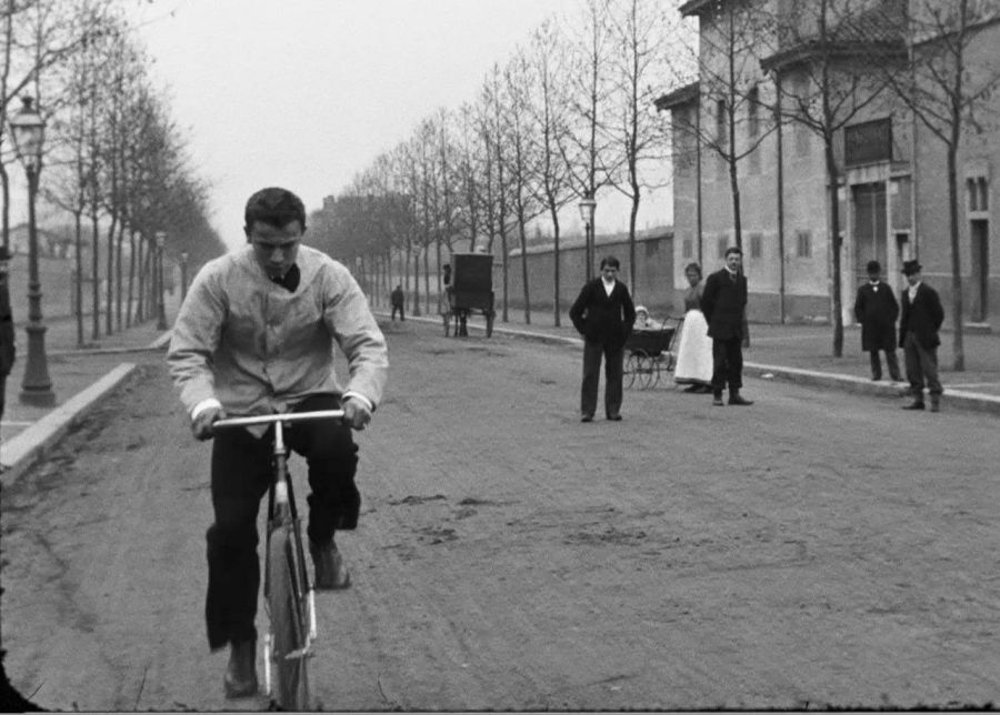 Escena callejera de principios del siglo XX: ciclista en bicicleta antigua, peatones, carruaje y cochecito.  Fotograma en blanco y negro.