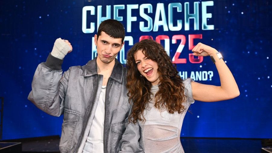 Dos personas, un hombre con chaqueta gris y una mano vendada y una mujer con camiseta gris clara, celebran con los brazos levantados.  Fondo con el logo 