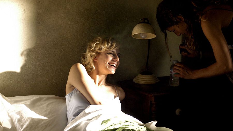 En una habitación iluminada por el sol, una mujer rubia sonríe en la cama mientras otra, de cabello oscuro, se acerca con una botella. Ambiente tranquilo.